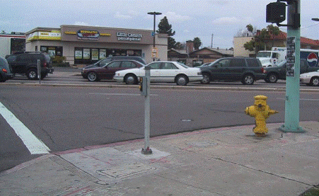 "Photo from sidewalk looking across street with street parallel to photographer on left. Pole and fire hydrant are on right in photo. Additional small pole, about 4 feet high, is located near the crosswalk line, about 2 feet from the curb; APS is installed on that pole.