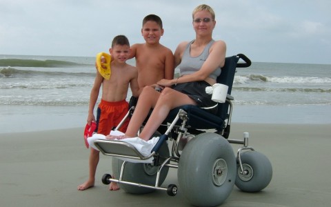 Woman using beach wheelchair with two young boys standing beside her