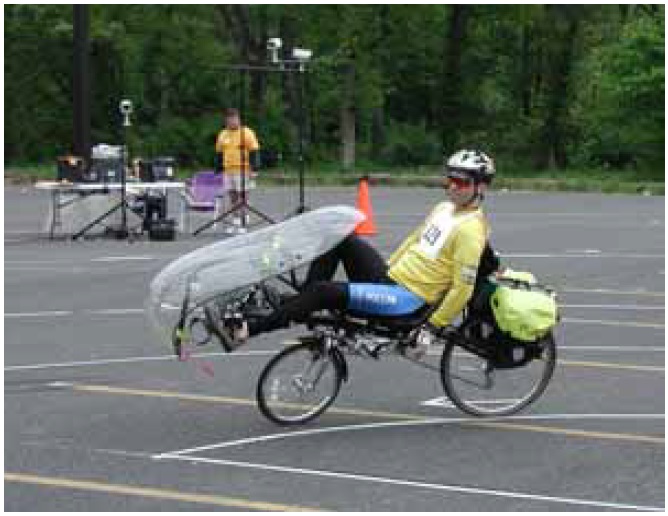 Figure 5: Photo. Recumbent bicycle. A man is riding a recumbent bicycle in a parking lot.