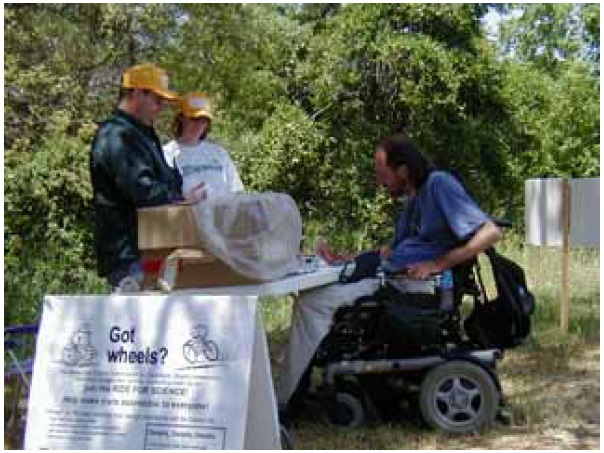 Figure 9: Photo. Power wheelchair. A man is in a power wheelchair. He is next to a table that has been set up alongside a trail.