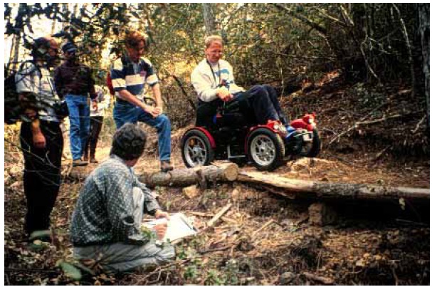 Figure 14: Photo. Off-road racing wheelchair. A man is in an off-road racing wheelchair. He is on a trail in the woods.