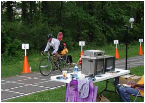 Figure 22: Photo. Video cameras were setup to record participant movements at Stations 3 through 7. A bicyclist is riding through the deceleration station. An event staff person adjacent to the trail is holding up a STOP sign. A video camera is mounted adjacent to the trail and is filming the bicyclist's reaction to the STOP sign.