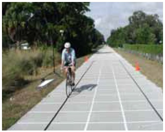 Figure 23: Photos. Equipment testing at data collection stations. This is a mosaic of five photos. 
Photo 23a: A bicyclist riding through the acceleration station. He is being filmed by a video camera mounted adjacent to the trail.