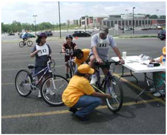 Figure 26: Photos. Physical measurements. Photo 26b: Two event staff persons are measuring the length of a bicycle and the width of the bicyclist. Two other bicyclists wait their turn.