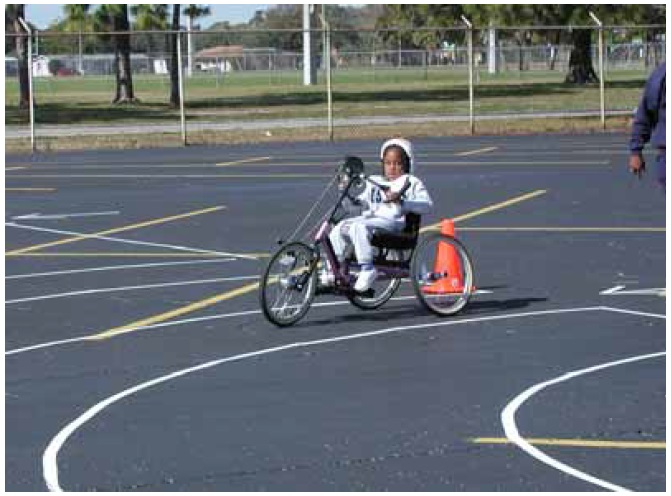 Figure 28: Photo. Participant within turning radii station. A participant is riding a hand cycle through a curved turning path delineated in a parking lot.