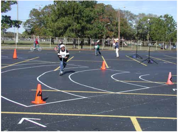 Figure 33: Photo. Participants traveling through progressively smaller turning radii. A participant on a nonmotorized kick scooter is traveling through the turning path with the second smallest radius. In the background, another participant on a nonmotorized kick scooter is approaching the turning path with the fourth smallest radius. They are riding on a black topped parking lot where lines have been placed to indicate the arc of the curve in which participants should travel. 