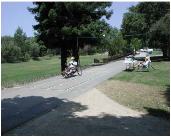 Figure 39: Photo. Speed (and sweep width) station. This is a photo of what actually took place as represented by the drawing in figure 38. We see a section of trail is marked with longitudinal lines spaced 1 meter apart. A participant on a hand cycle is travels through this station. Transverse lines mark the beginning and end of the sweep width and speed station. An overhead video camera is films him. An event staff person is sits at a table and observes the participant.