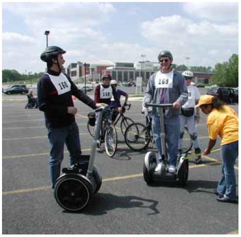 Figure 46: Photo. Trail users have diverse operating characteristics. An event staff person is taking measurements of a participant standing on a Segway. Another participant beside a Segway, a participant using inline skates, and two participants on bicycles are waiting to be measured.