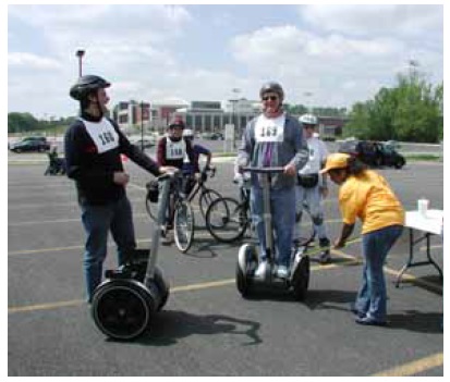 Figure 50: Photo. Segway users at the physical measurements station. An event staff person is taking measurements of a participant standing on a Segway. Another participant beside a Segway, a participant using inline skates, and two participants on bicycles are waiting to be measured.