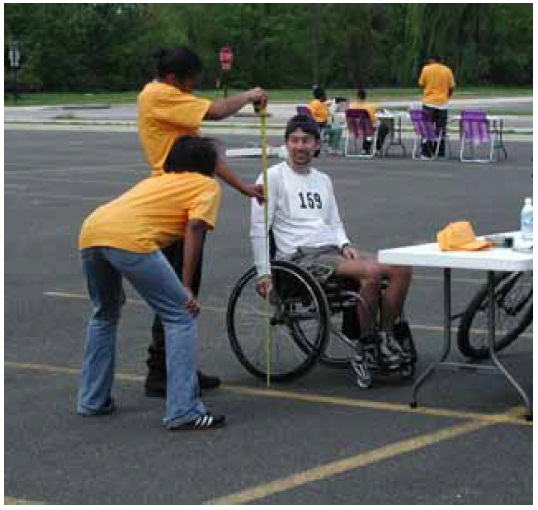 Figure 26: Photos. Physical measurements. Photo 26a: Two event staff persons are measuring the height of a participant in a manual wheelchair.