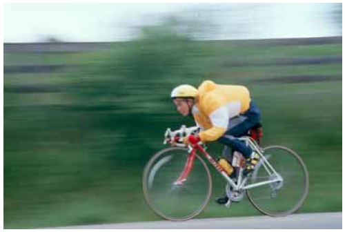 Figure 47: Photo. AASHTO's design bicyclist travels at 30 kilometers/hour (20 miles/hour). A bicyclist is riding at a high rate of speed as indicated by the blurred green background behind him and his wind-filled jacket.