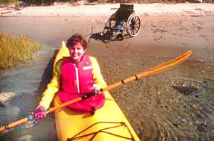 photo of a woman in a kayak In the water, with a wheelchair in the background on a sandy beach