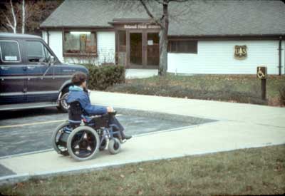 Image of a person in a motorized wheelchair moving up a sidewalk towards a building.