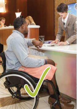 Man in a wheelchair checking in at a lowered hotel check in counter with a green check mark at the bottom of the image, indicating an accessible element