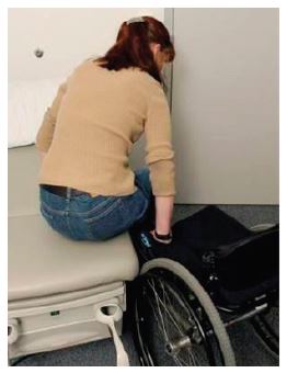 A woman moves herself from an adjustable height examining table to her wheelchair