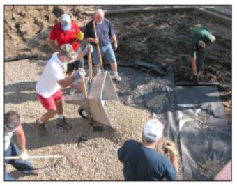 group of people working to spread rocks on draingage fabric, with one man overturning a wheelbarrow of rock and several other men spreading the rock wit rakes