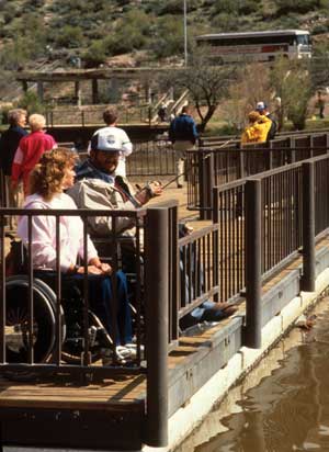 Photo of a man and woman chatting as the man fishes from a dock with lowered guard rail to allow easier fishing while using a wheelchair.