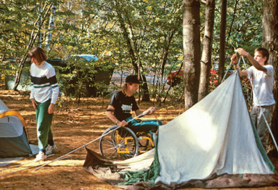 Photo of three people setting up a tent. One of the individuals is using a wheelchair.