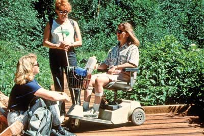 Photo of three people talking on a boardwalk. One of the individuals is using a motorized scooter-type wheelchair. Another is holding a probing white cane.