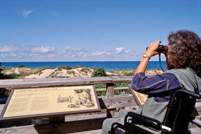 Photo of a person using a wheelchair at an interpretive station. The sign, mounted on a table-like metal stand, is situated at a level that can be read by a person using a wheelchair. The woman is using binoculars to look over the top of the interpretive signs at the dunes and ocean.