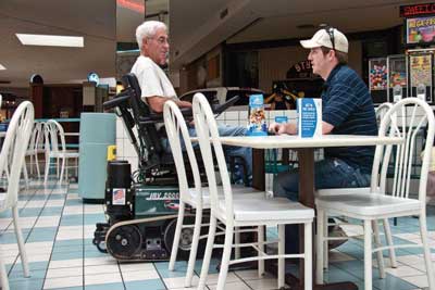 Photo of two men chatting at a table in a mall food court.  One man is using a wheelchair that moves using smooth rubber belts rather than wheels.
