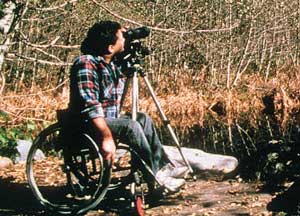 Photo of a man using a "standard" wheelchair at an informal viewing area by a lake in a forest.