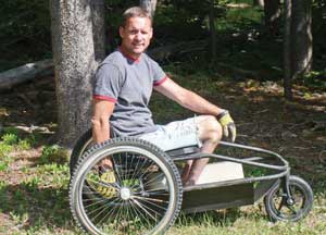Photo of a man using a low, external frame off-road wheelchair with two large rear wheels and a smaller front wheel, in a forest.
