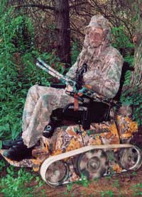 Photo of a man wearing camouflage hunting clothing and carrying a crossbow, using a belt-track type wheelchair that has been camouflage painted.