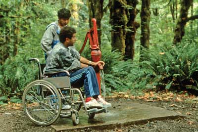 Photo of two people at a handpump in a forested area. One person is using a wheelchair. The other person is pumping water using the long handled pump.
