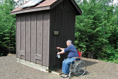 Photo of a man beside a small building filling a water bottle from a spigot.  The building has two solar panels on the roof.