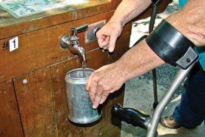 Photo of a man filling a coffee pot with water from a spigot on the side of a wooden box that also supports a drinking fountain.  A solar panel is mounted on top of a tall steel pole behind the man.