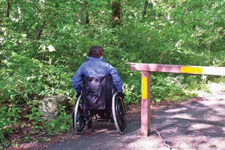 Photo of a woman who is using a wheelchair going around a steel road closure gate on a paved road. The space between the gate end post a large boulder is sufficient for passage by a person using a wheelchair, but not a vehicle.  This bypass is also flat and the surface is paved, allowing easy passage.