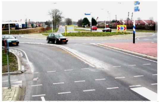 image of two cars separated by a traffic island, exiting a roundabout approaching pedestrian and bicycle crossings.