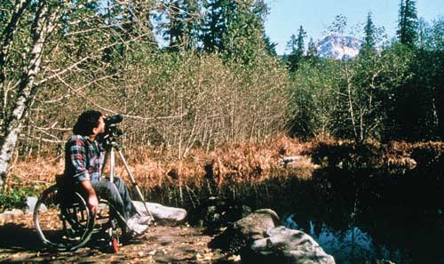 Photo of a man using a wheelchair beside a lake. He is looking through a tripod-mounted camera with a telephoto lens at a snow-covered mountain in the background.
