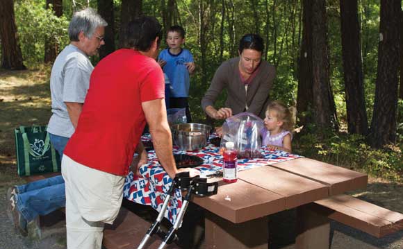 Photo of people of three generations enjoying a picnic lunch at a table in a forested area.  One of the people uses crutches.