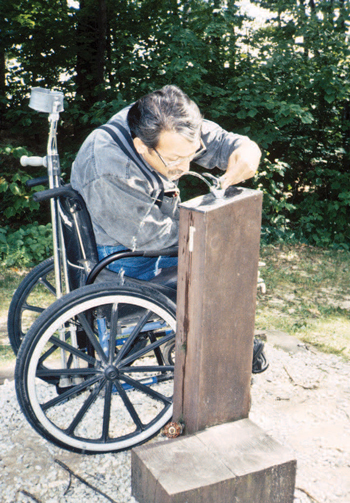 Photo of a man using a wheelchair leaning over to drink out of a fountain that is mounted on a wood post in a forested area.