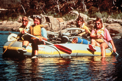 Photo of four people rafting on a river. The raft contains camping equipment and a wheelchair.