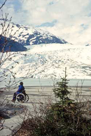 Photo of a person in a wheelchair on a paved path looking across a lake at a glacier.