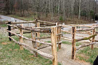 Photo of a chicane made from log fence poles at the edge of a field near a road.  The chicane is an “S” shaped pathway through a fence that allows people to pass but is sized so that large farm animals and motorized vehicles are too large to fit through the multiple corners that comprise the “S” shape.