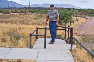 Photo of a man opening a kissing gate to access a concrete walkway.  The kissing gate consists of a square enclosure with two adjacent openings that lead to opposite sides of a fence.  The gate swings so that only one opening can be accessed at a time.  This gate is made of steel tubing and is sized so that people can pass but the enclosure is too small to allow the passage of motorized vehicles.