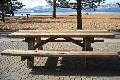 Photo of a wood picnic table on an accessible paving stone surface overlooking Lake Tahoe and a sand beach.  Pine trees are scattered about the area and mountains are visible in the distance.