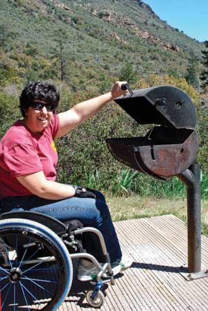 Photo of a woman who uses a wheelchair opening the lid of a pedestal grill shaped like a small barrel laid on its side that is elevated and cantilevered from a steel post.