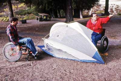 Photo of two men pitching a small tent on a coarse sand surface in a forested campground.  Both men are using wheelchairs.