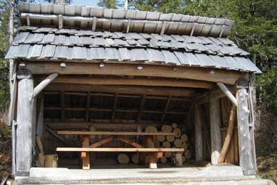 Photo of a person walking into a rustic camp shelter made of cedar logs, with a shingle roof.  The shelter seems tiny compared to the huge cedar and fir trees around it.