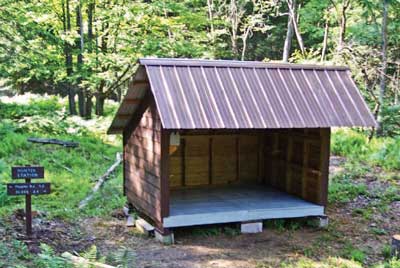 Photo of a small, frame camp shelter with a floor that is raised above the ground, shingled walls, and a metal roof.  Near the shelter, there is a bench and a wood sign stating “Hunter Station” and giving distances to destinations to the right and left down a trail.