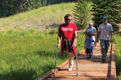 Photo of people of three generations enjoying a stroll on a boardwalk in a meadow.  One of the people uses crutches.