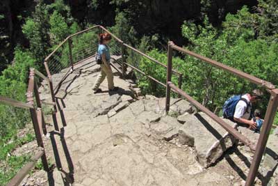 Photo of a trail section consisting of steps chiseled into a natural rock cliff.  Metal handrails have been provided for the hikers.  The vertical drop of the section of trail appears to be about 50 feet, but it is much further to the valley below.