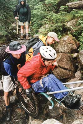 Photo of 4 people on a very rocky trail.  Two people are assisting by a hiker in a wheelchair up a cliff-like section of the trail by pushing from behind. Pulling poles are hooked to the front of the wheelchair so that other people who are not in the photo can also assist by pulling from the front of the wheelchair.