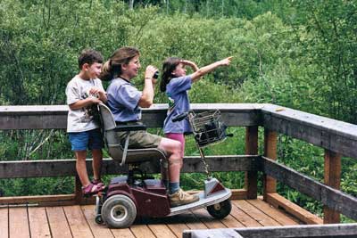 Photo of a mother and her two children watching waterfowl from an overlook at a lake.  The mother is using a motorized scooter.
