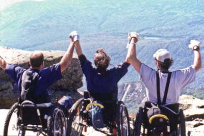 Photo of three people who use wheelchairs with their hands joined in victory above their heads, on a rock outcrop overlooking a forest. 